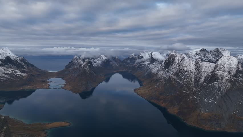 Aerial view of snow-capped Reine Mountains reflected in the serene waters, creating a stunning contrast of textures, Reine, Nordland, Norway.