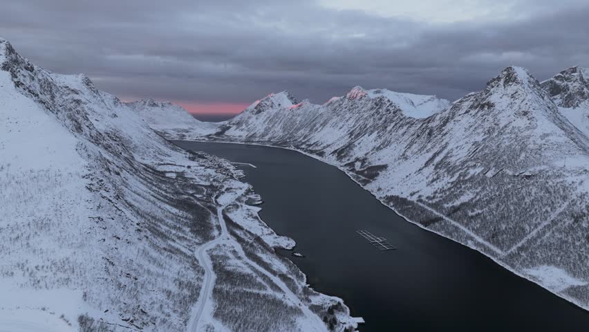 Aerial view of snow-covered mountains, a winding road, and dark waters creating a striking contrast against the dusky sky, Gryllefjord, Troms, Norway.