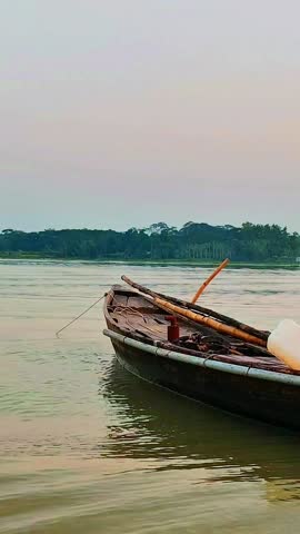A lone wooden boat drifts and gently rocks on calm river waves during golden hour. Serene Bangladeshi river scene with warm sunset light reflecting on water. 