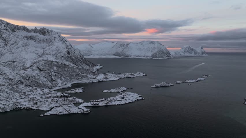 Aerial view of snow-blanketed mountains meeting the dark ocean, dotted with islands under a cloudy sky, Gryllefjord, Troms, Norway.