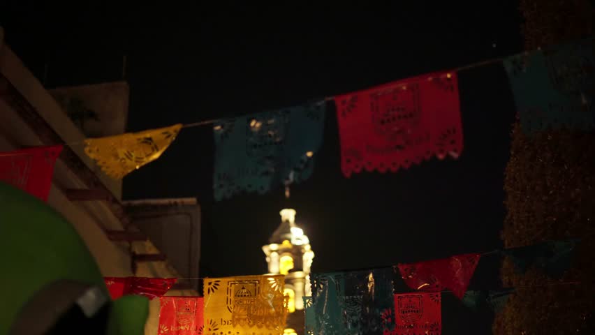 Colorful papel picado hang across a dimly lit street at night, swaying above warm archway light while a softly illuminated tower rises in the background, creating a festive and atmospheric scene