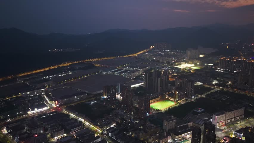 Aerial night view of Shenzhen’s Pingshan District, highlighting the illuminated BYD headquarters, glowing city lights, sports fields, and distant mountains in a vibrant urban scene.