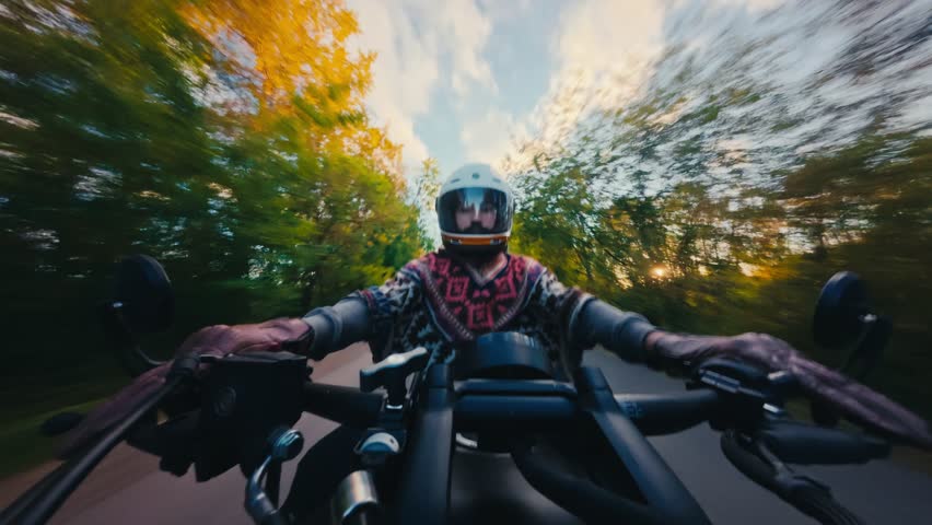 Motorcyclist riding a bike on a forest road captured by a selfie camera. Rider wearing a helmet with a gold visor and a bomber jacket driving fast.