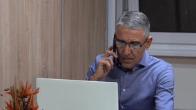 Middle-Aged Man Takes Phone Call While Working on Laptop at Home - Zoom In Shot. During the call he pointing at the computer screen. Perfect for remote work, communication, and professional themes. - Powered by Shutterstock - Get 15% off with code: PIKWIZARD15