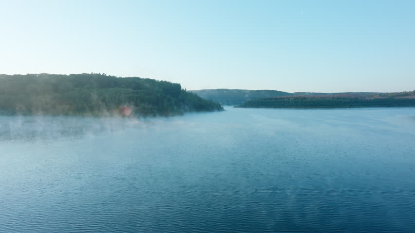 High quality video showing a calm lake at dawn with soft mist drifting across the water and gentle morning light over distant hills.