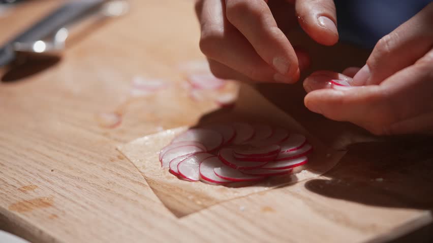 A person is cutting radishes on a wooden cutting board. The radishes are sliced into small pieces and are placed on a plastic wrap