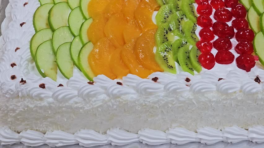A rectangular white cream cake topped with rows of colorful fresh fruit slices, including kiwi, peach, and red berries, with a coconut flake side coating.