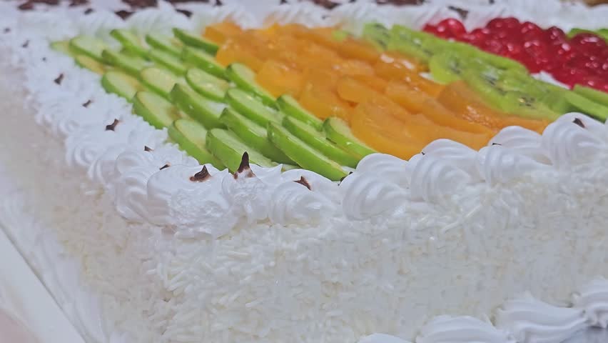 A rectangular white cream cake topped with rows of colorful fresh fruit slices, including kiwi, peach, and red berries, with a coconut flake side coating.