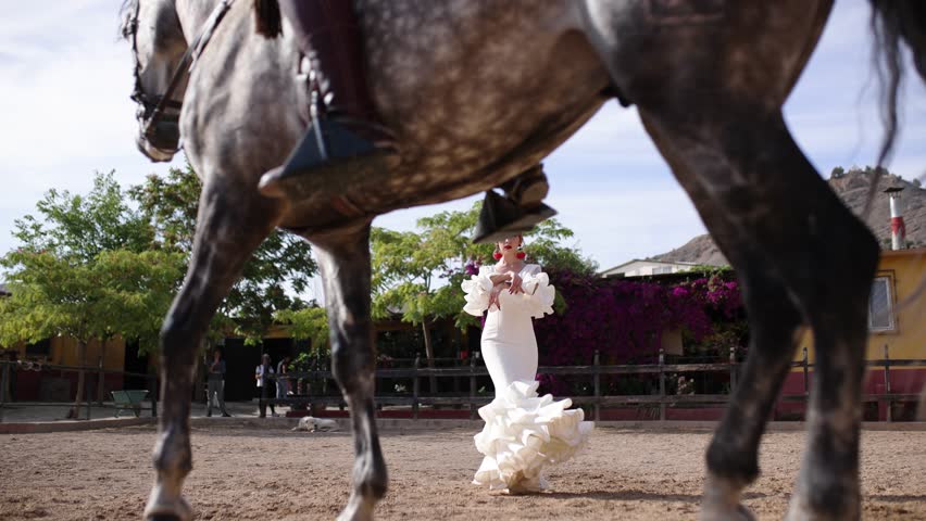 Elegant woman in a traditional white flamenco dress dancing with passion in a sandy arena. A beautiful dapple gray horse passes in front of her