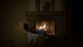 A girl sits in an armchair with her back to the camera, warming her feet in cozy knitted socks. The burning fireplace is centered in the frame, casting a warm, cozy glow across the room - Powered by Shutterstock - Get 15% off with code: PIKWIZARD15