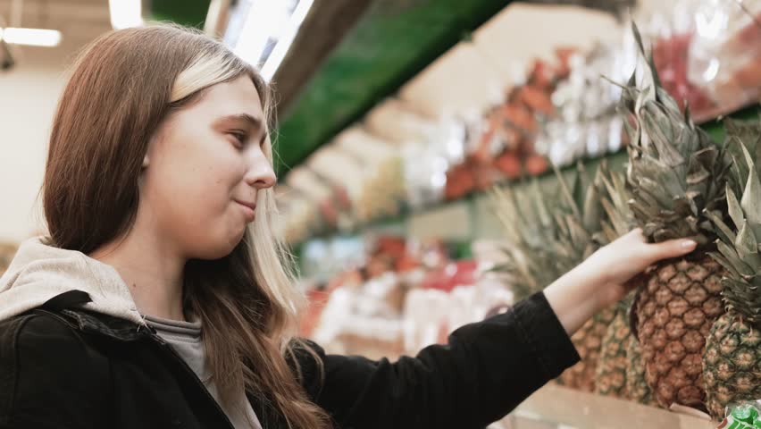 A girl in a black jacket with long hair chooses pineapples in a hypermarket.
