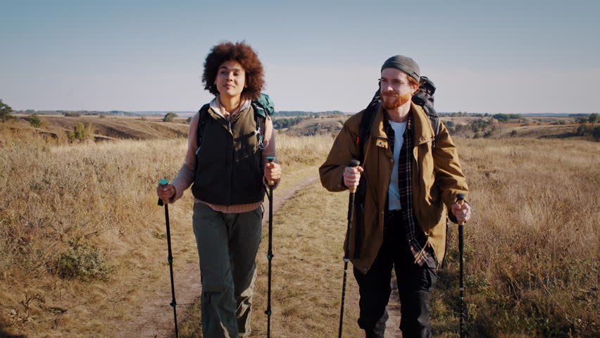 Red bearded man with black girlfriend engages in Nordic walking at autumn countryside slow motion. Young sportive family hikes on hilly rural site on sunny day