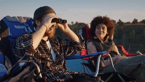 Young red-bearded man gives binoculars to black woman to observe horizon as rest at tents camp. Mixed race family enjoys calm evening after active trekking day - Powered by Shutterstock - Get 15% off with code: PIKWIZARD15