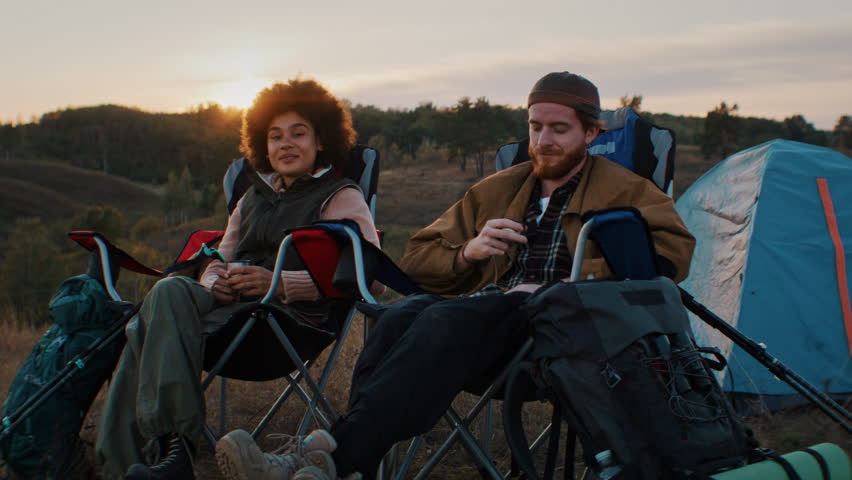 Happy tired tourists drink tea while sit on folding chairs at evening camp. Young mixed race couple rests refreshing with hot beverages at sunset countryside
