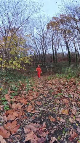 A man in red jumps through leaves amid autumn forest with bare trees and overcast sky, dynamic midair splits and kicks captured in motion, energetic martial artist and athlete demonstrates flexibility