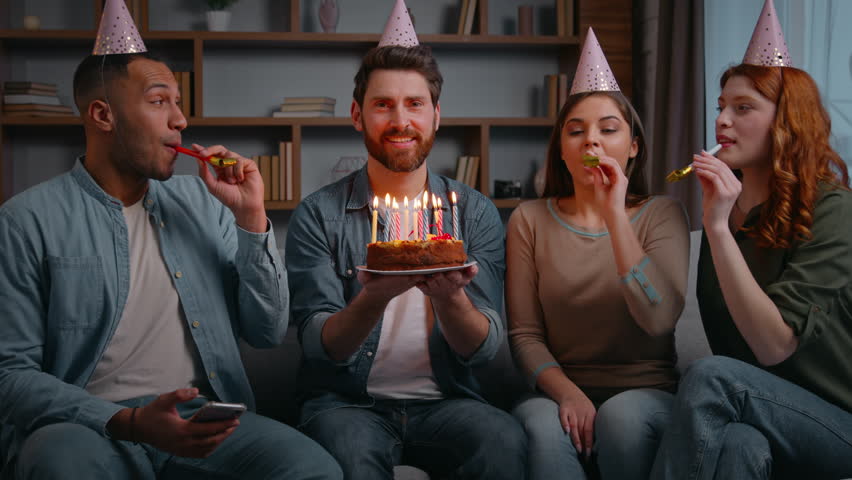 Four multiracial cheerful friends at home party congratulate birthday man laughing carefree smiling holding cake with candles happy diverse group men women surprise event celebration festive meeting 