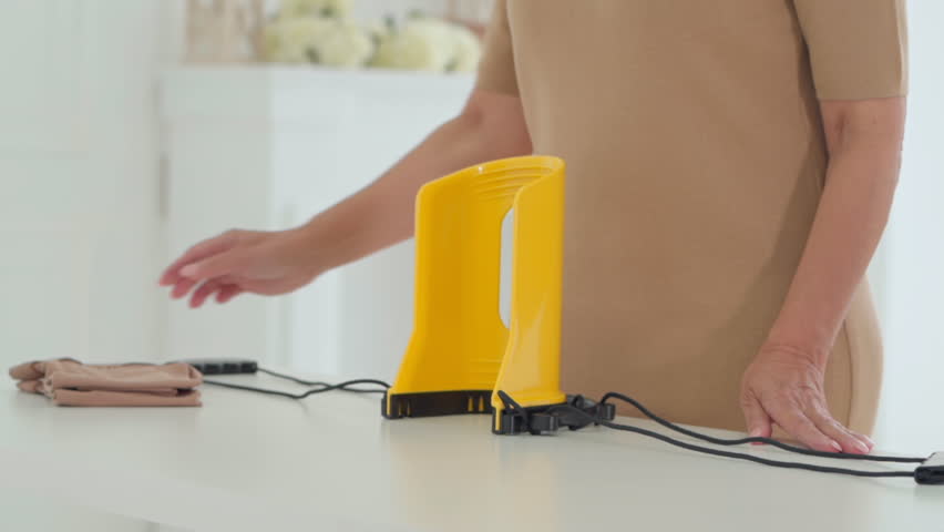 A senior woman places a beige compression stocking onto a yellow donning aid, showing the simple preparation process for wearing medical support hosiery at home.