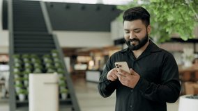 Portrait of happy Indian businessman uses smartphone in office building lobby. Young bearded entrepreneur scrolls internet on mobile phone in coworking. Communication technology - Powered by Shutterstock - Get 15% off with code: PIKWIZARD15