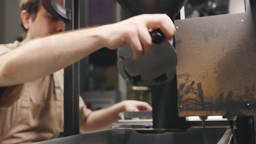 Freshly roasted coffee beans are being ground in a roasting facility. A skilled worker closely monitors the process, ensuring optimal flavor extraction.