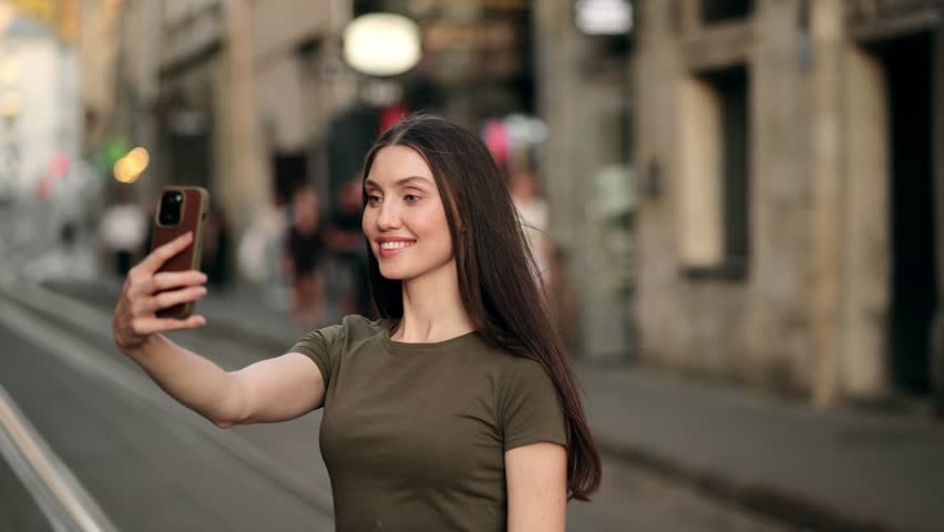 Young woman taking a selfie on a city street at sunset, adjusting her hair and posing with a smartphone. Urban background with soft bokeh lights creates a warm and stylish atmosphere
