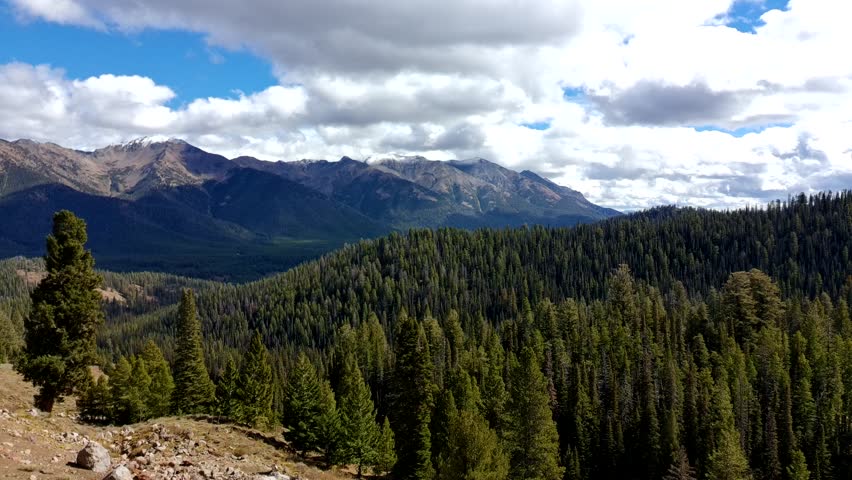 A mountain range with a cloudy sky and a forest below. The sky is blue and the mountains are covered in trees