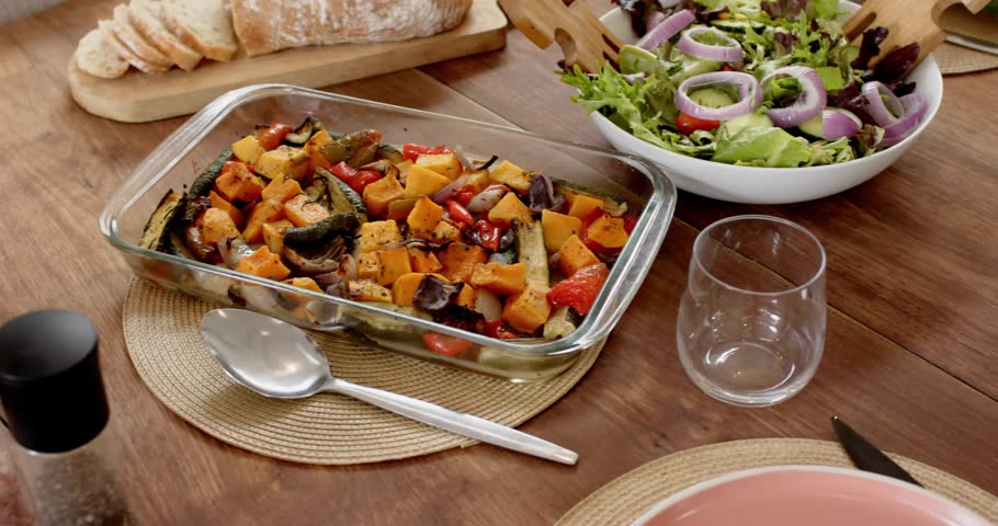 Camera starting slow pan revealing salad bowl, roasted veg, bread, chicken, showing table set. Greens, dish, platter, loaf, onion, placemats, glassware