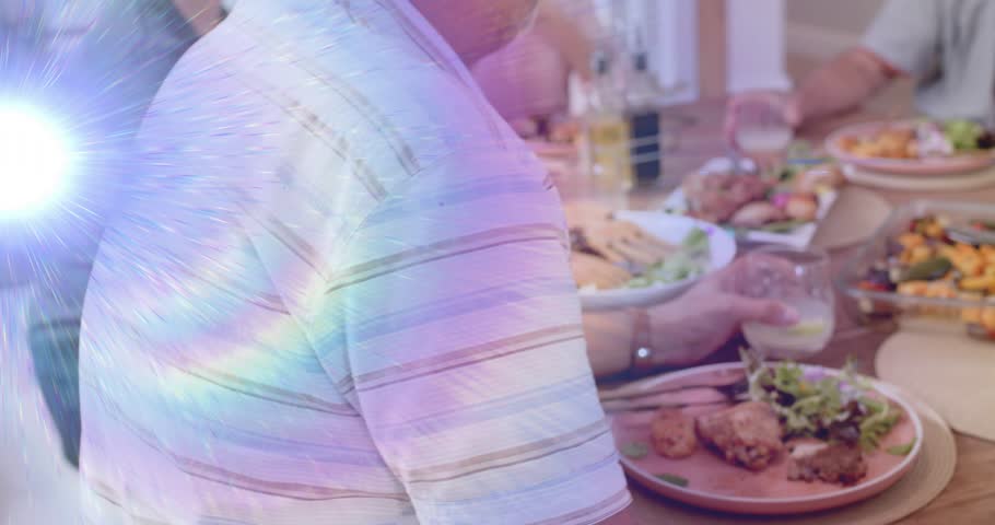 Man turning toward diners after starting toast, raising stemless glass amid spectral lens flare. Family, dining, meal, home, wooden, table, convivial