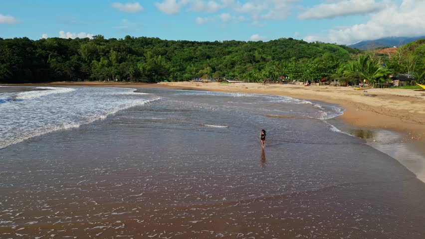 Ground‑level aerial tracking a woman running along the sandy shoreline, framed by gentle waves, palm trees, and lush tropical scenery in Quinawan Beach, Mariveles, Bataan, Philippines.