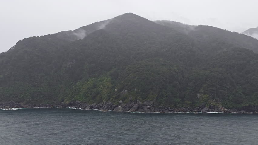 Milford Sound in New Zealand presenting towering, verdant mountains descending into the dark waters of the fjord, enveloped in atmospheric clouds and mist on cloudy day