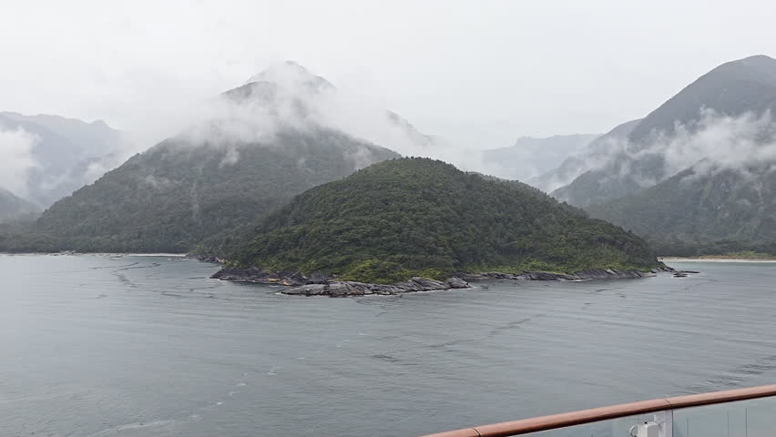 Milford Sound in New Zealand presenting vast fiord with an island covered in native forest, surrounded by towering, fog shrouded peaks on cloudy day