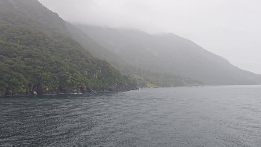 Milford Sound fiord during cloudy and rainy day. Displaying dramatic towering mountains shrouded in mist. With lush green rainforest clinging to the steep coastline and dark water below