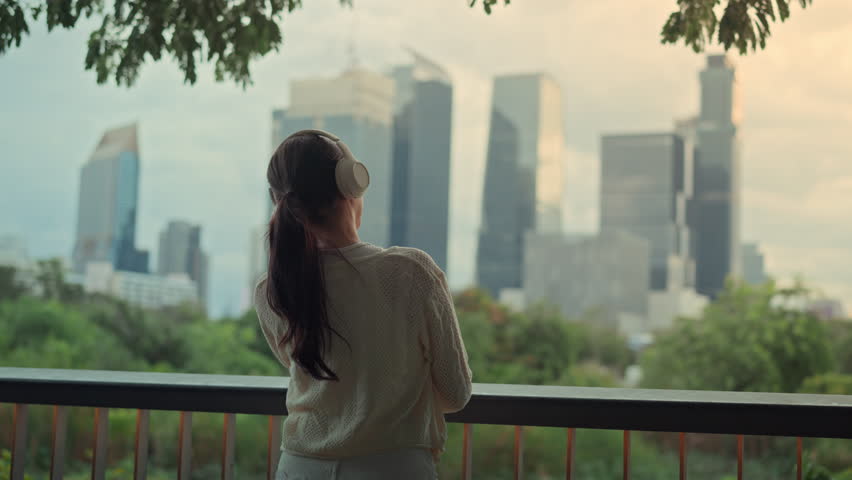 Asian woman listening to music with headphones while looking at the city skyline, relaxing on balcony, urban lifestyle, peaceful morning mood, modern city living and mindfulness moment