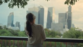 Asian woman listening to music with headphones while looking at the city skyline, relaxing on balcony, urban lifestyle, peaceful morning mood, modern city living and mindfulness moment - Powered by Shutterstock - Get 15% off with code: PIKWIZARD15