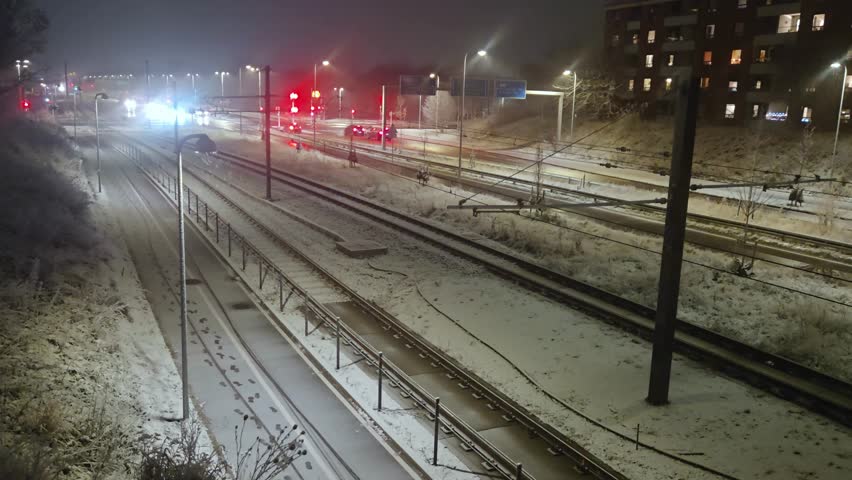 Denmark, Vallensbaek Strand  winter - A winter night scene captures snow-covered train tracks and a road with illuminated cars, streetlights, and a multi-story building under a dark sky.