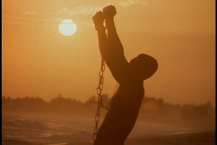 Slavery reenactment in Ghana, Africa. MS silhouette of black man in chains crouching on beach. He stands and raises his arms. | Shutterstock HD Video #3947549