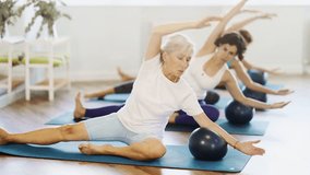 Concentrated young adult Hispanic woman, fitness enthusiast, participating group Pilates session in cozy sunlit studio, doing side bend with exercise ball, promoting balance and lateral stretching - Powered by Shutterstock - Get 15% off with code: PIKWIZARD15
