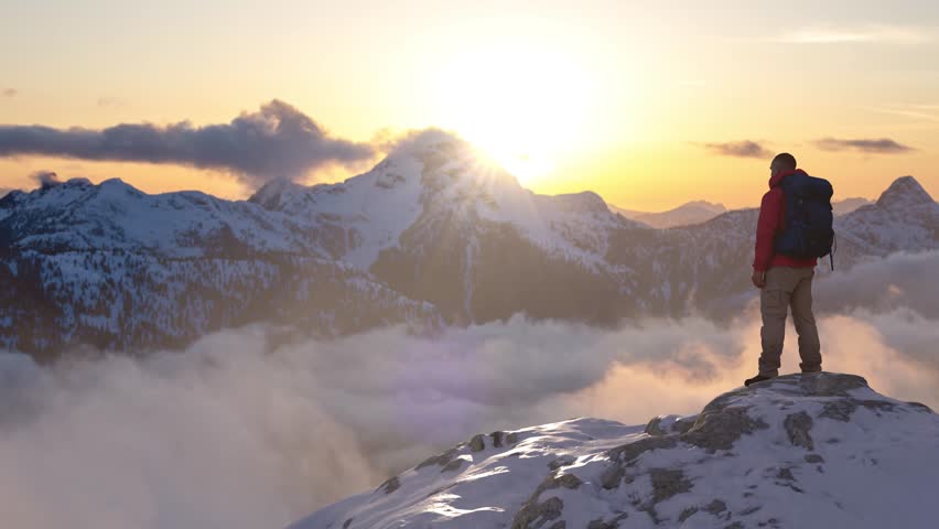 Adventurer on a Snow-Covered Mountain Peak Gazing at a Golden Sunrise Over Cloudy Peaks