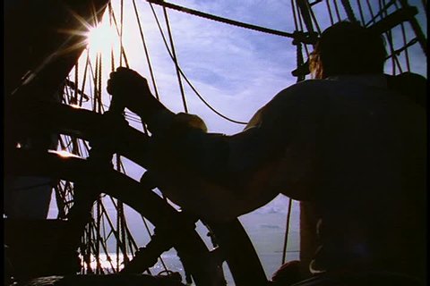 Historical reenactment of HMS Bounty ship on Rhode Island. MS silhouette of crew member with hand on ship's wheel. Rigging, open sea, clouds and blue sky in background.