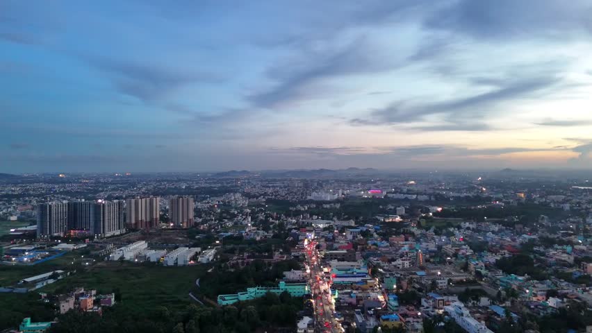 Chennai’s cityscape at sunrise shows dense older streets in the foreground and tall apartment towers. Streetlights and traffic trails brighten the scene, with distant hills visible on the horizon.
