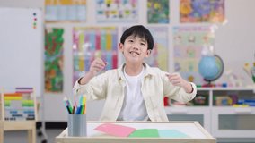 Smiling boy raises fists in excitement, sitting at desk in bright, colorful classroom environment - Powered by Shutterstock - Get 15% off with code: PIKWIZARD15