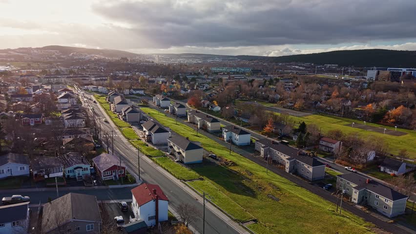 Aerial view of a suburban street lined with neat houses, green lawns and autumn trees, stretching toward distant hills under a partly cloudy sky.