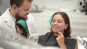 Interested young woman sits in a dentist chair and carefully looks at her teeth in the mirror. Male dentist with an assistant shows the client the work done. High quality 4k footage - Powered by Shutterstock - Get 15% off with code: PIKWIZARD15
