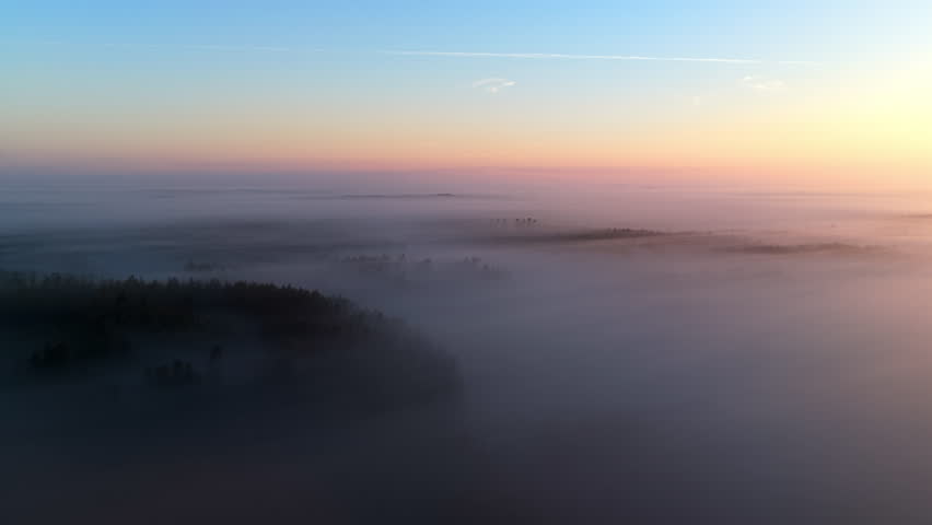Aerial tracking shot overlooking foggy fields covering gloomy forest, sunrise
