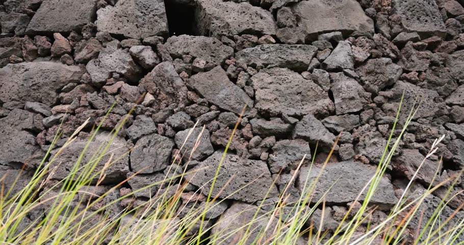 A wall made of volcanic rocks and a grassy area in front of it. The wall is made of large rocks and the grass is short. Trucking shot.