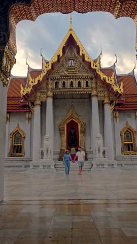 A couple stands hand-in-hand in front of the stunning Wat Benchamabophit in Bangkok, Thailand. The ornate architecture and golden details create a romantic atmosphere during sunset.