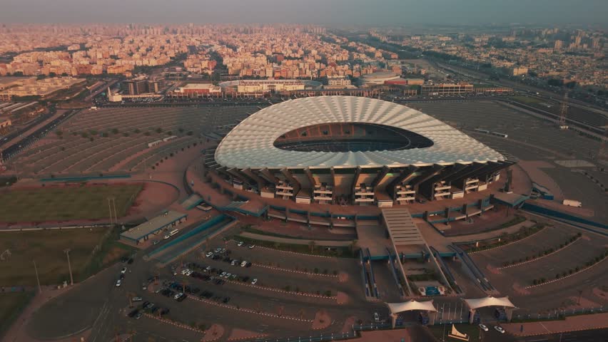 A cinematic 4K drone shot capturing Jaber Al-Ahmad International Stadium during sunrise in Kuwait City. The footage highlights the modern architectural roof, the vast surrounding infrastructure,