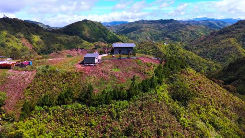 drone shot captures several modern homes perched on a grassy hilltop amid rolling, tree‑covered mountains. A winding road leads up to the houses, the scene highlights verdant landscape.The bright sky