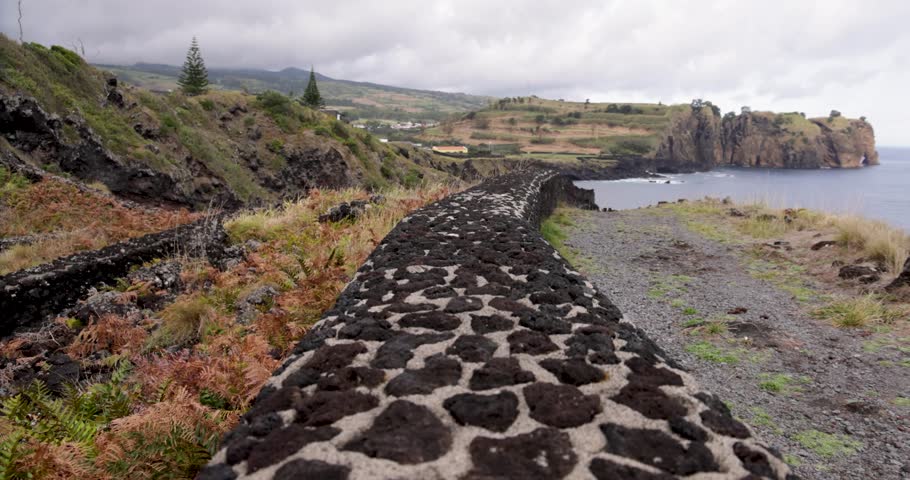 Walking on a wall at a rugged shoreline with large rock formations in a cove at Sao Vicente Ferreira, Azores. The water is calm and the sky is cloudy. Tracking shot.