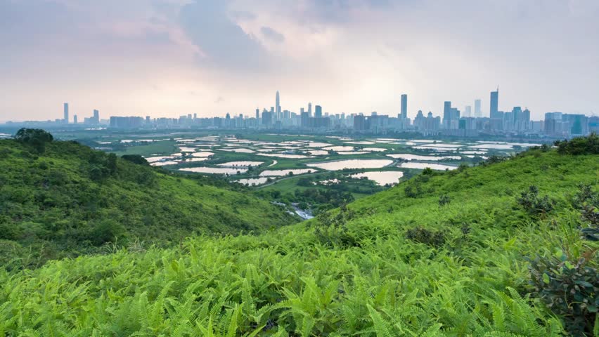 Lush green hills overlook a distant cityscape with ponds under a cloudy sky