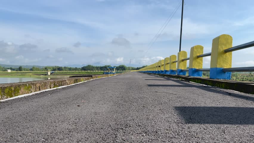 Roadside railing beside a tropical reservoir under bright morning skies, showing open landscape and calm weather. Low angle FPV move forward footage.